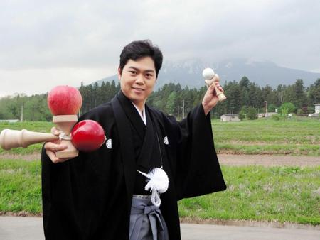 　岩木山をバックに、りんごを乗せたけん玉でポーズを決める三山ひろし＝青森県弘前市