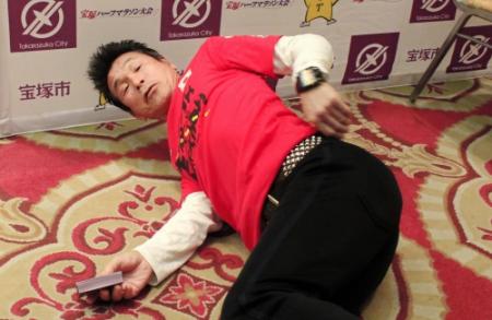 「宝塚大使」に任命された後、会見場で寝そべり座骨神経痛について説明する間寛平＝兵庫県宝塚市内