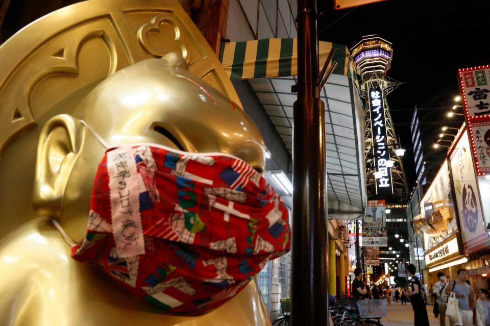 　大阪・新世界でマスクを着けたビリケンさん。奥は黄色にライトアップされた通天閣