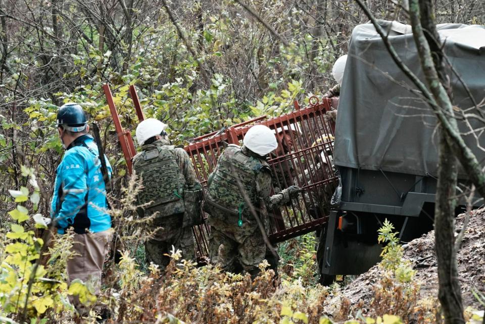 　銃を持ったハンター（左端）が警戒するなか、クマ対策の箱わなを運ぶ陸上自衛隊員ら＝秋田県鹿角市
