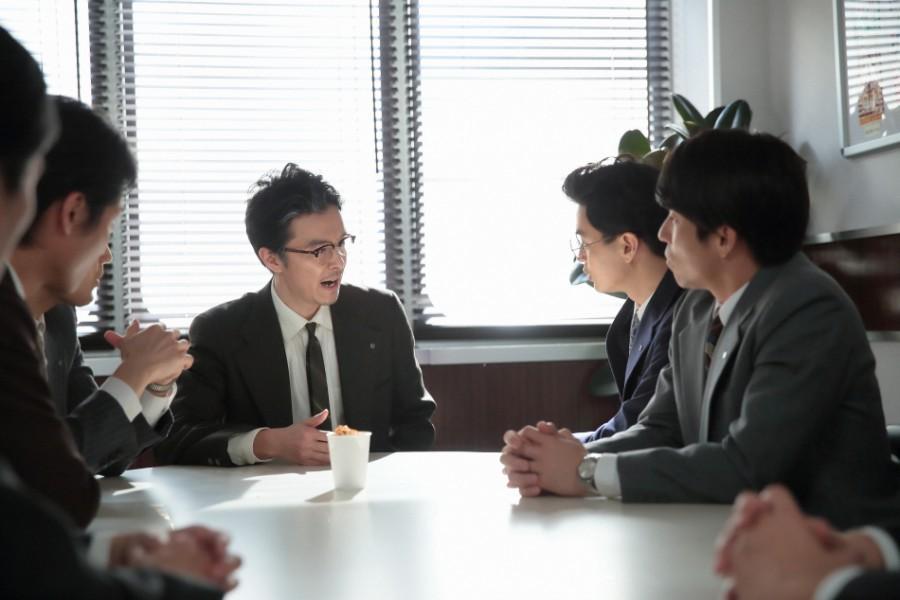 まんぷく食品の会議室で、割ったまんぷくラーメンを入れた紙コップを見せ、「容器に入ったラーメンを開発する」と社員に話す萬平（長谷川博己）