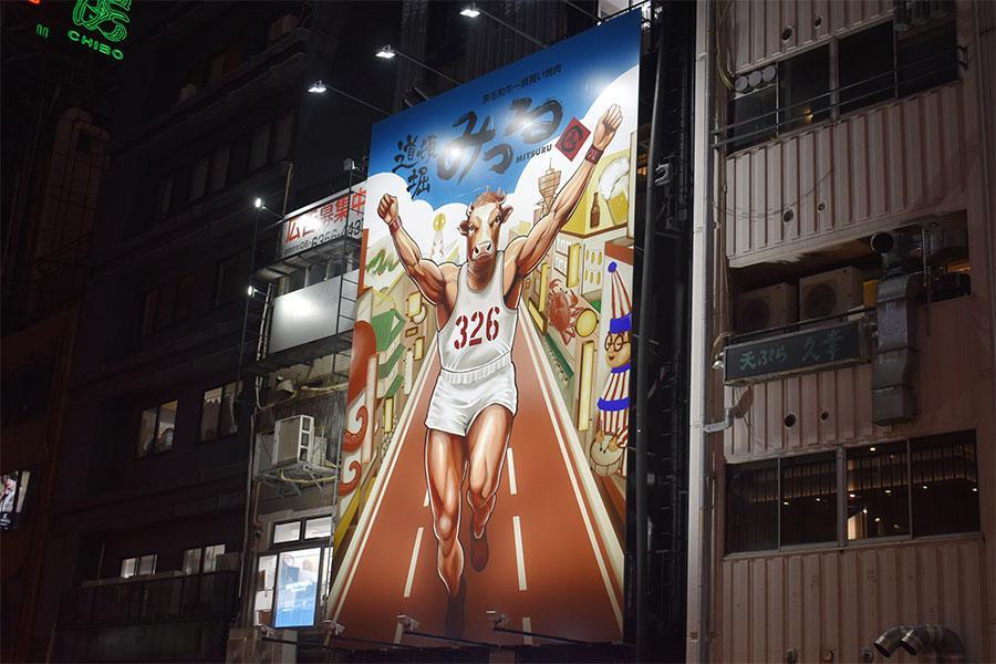 　新たに誕生した「黒毛和牛一頭買い焼肉 道頓堀みつる 本店」高さ１１mの巨大看板。グリコポーズの牛以外に、くいだおれ太郎や通天閣など大阪らしいスポットも描かれている