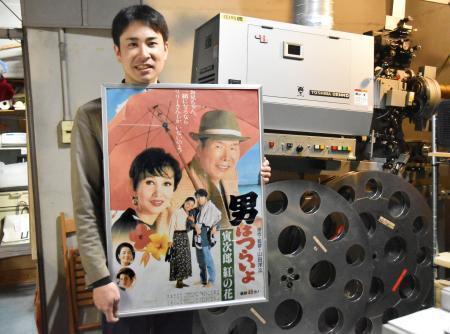 　上映に使うフィルム映写機の前で映画「男はつらいよ」のポスターを手にする小山岳志さん＝２３日、神戸市兵庫区