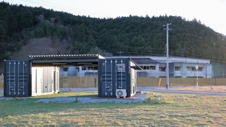 　震災遺構となった大川小の校舎（奥）北側の空き地に立つ「Ｔｅａｍ大川　未来を拓くネットワーク」のコンテナ事務所＝１０日午後、宮城県石巻市