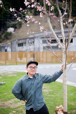 　大川小の校舎近くに咲いた桜と、木を植えた只野哲也さん＝１６日午後、宮城県石巻市