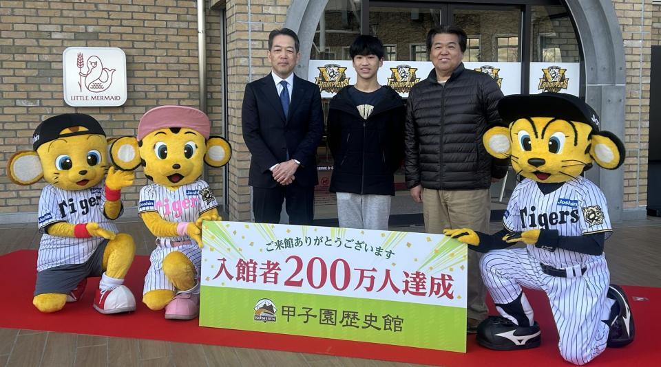 甲子園歴史館の入館者２００万人を達成し、上戸健司館長（左）やマスコットから祝福される田浦さん親子