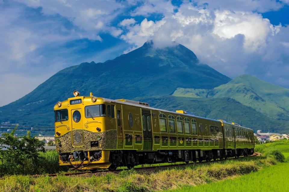 　由布岳をバックに快走する「或る列車」。金色に輝く車体と豪華な内装とスイーツも自慢だ（ＪＲ九州提供）