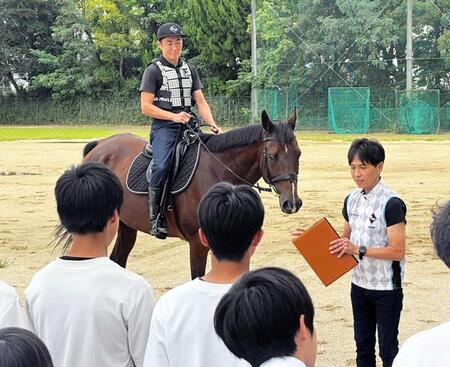 　運動場で常歩などを実演した馬上の佐久間助手と、渡辺師の話に熱心に耳を傾ける生徒たち＝滋賀県草津市立草津中学校